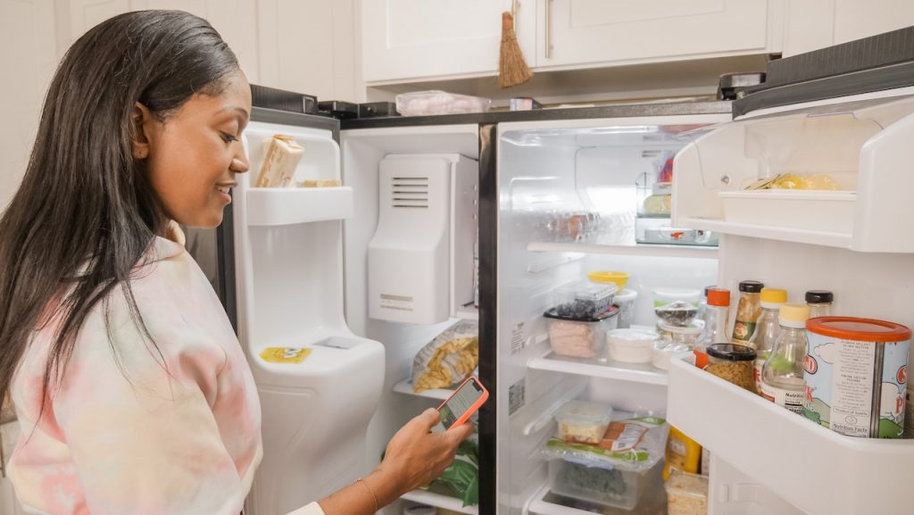 Woman standing in front of fridge with both the fridge and freezer doors open making a grocery list.