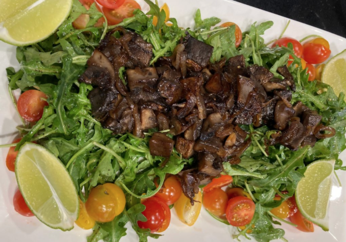 A bed of arugula lettuce with marinated mushrooms on top with cherry tomatoes and lime wedges